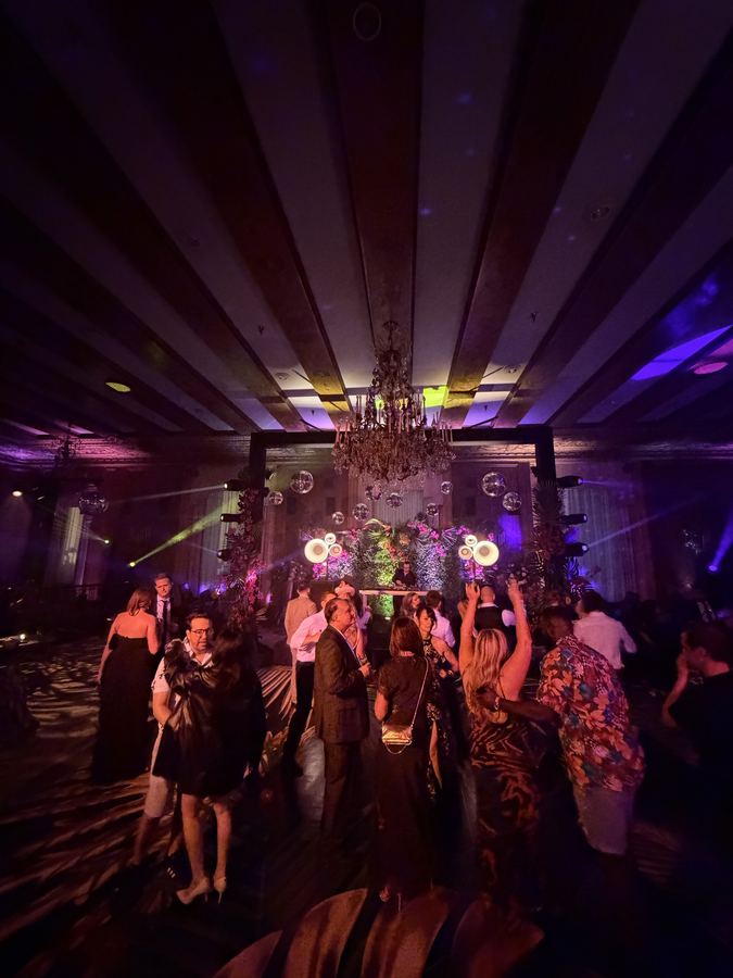 Guests in formal wear dancing in a ballroom with the stage and chandelier behind