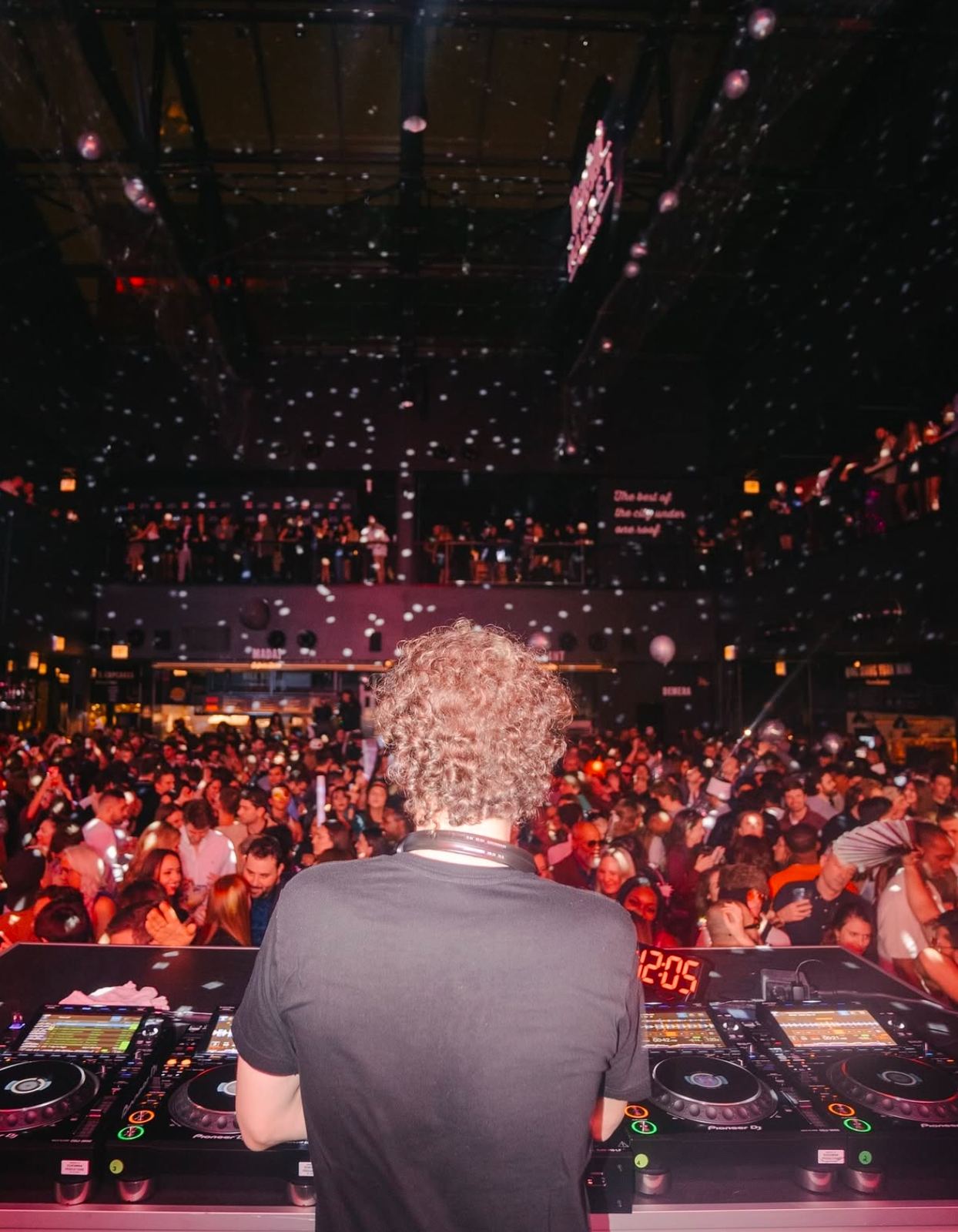 View from behind the DJ at the decks facing a packed crowd in a market hall