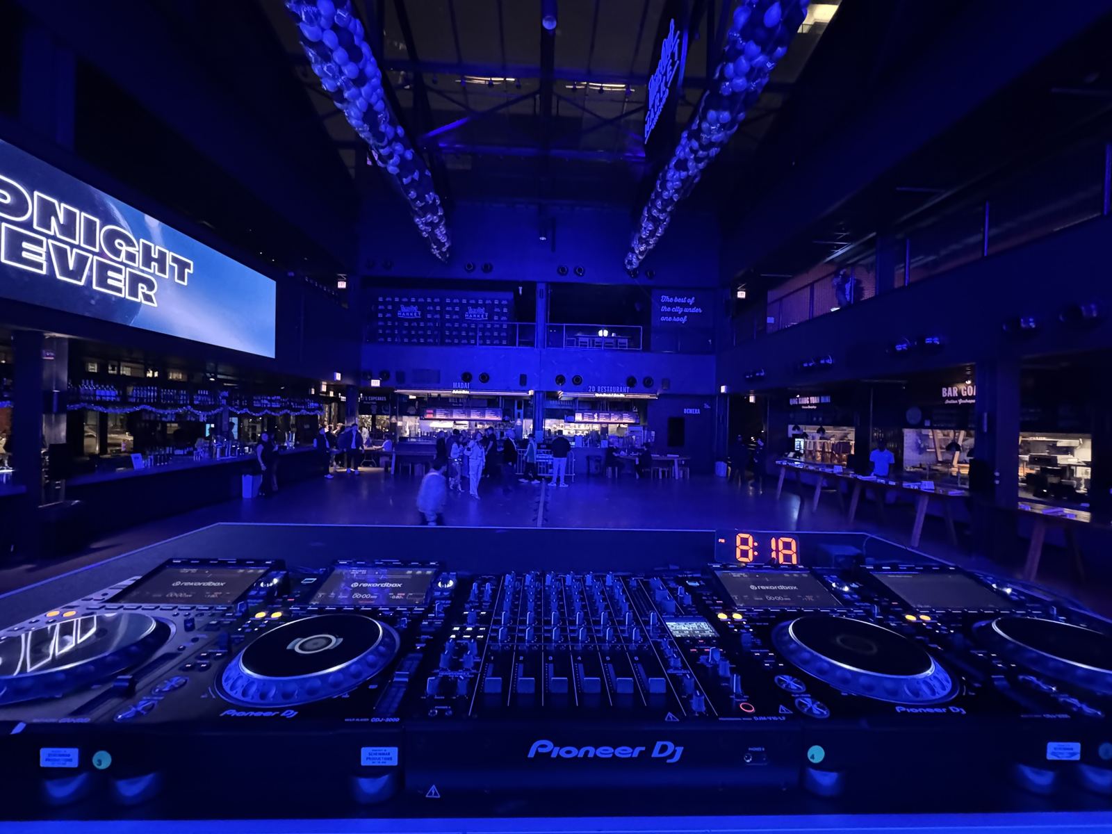 DJ booth with four CDJs and a Pioneer DJM-V10-LF mixer looking out over an empty market hall lit in blue